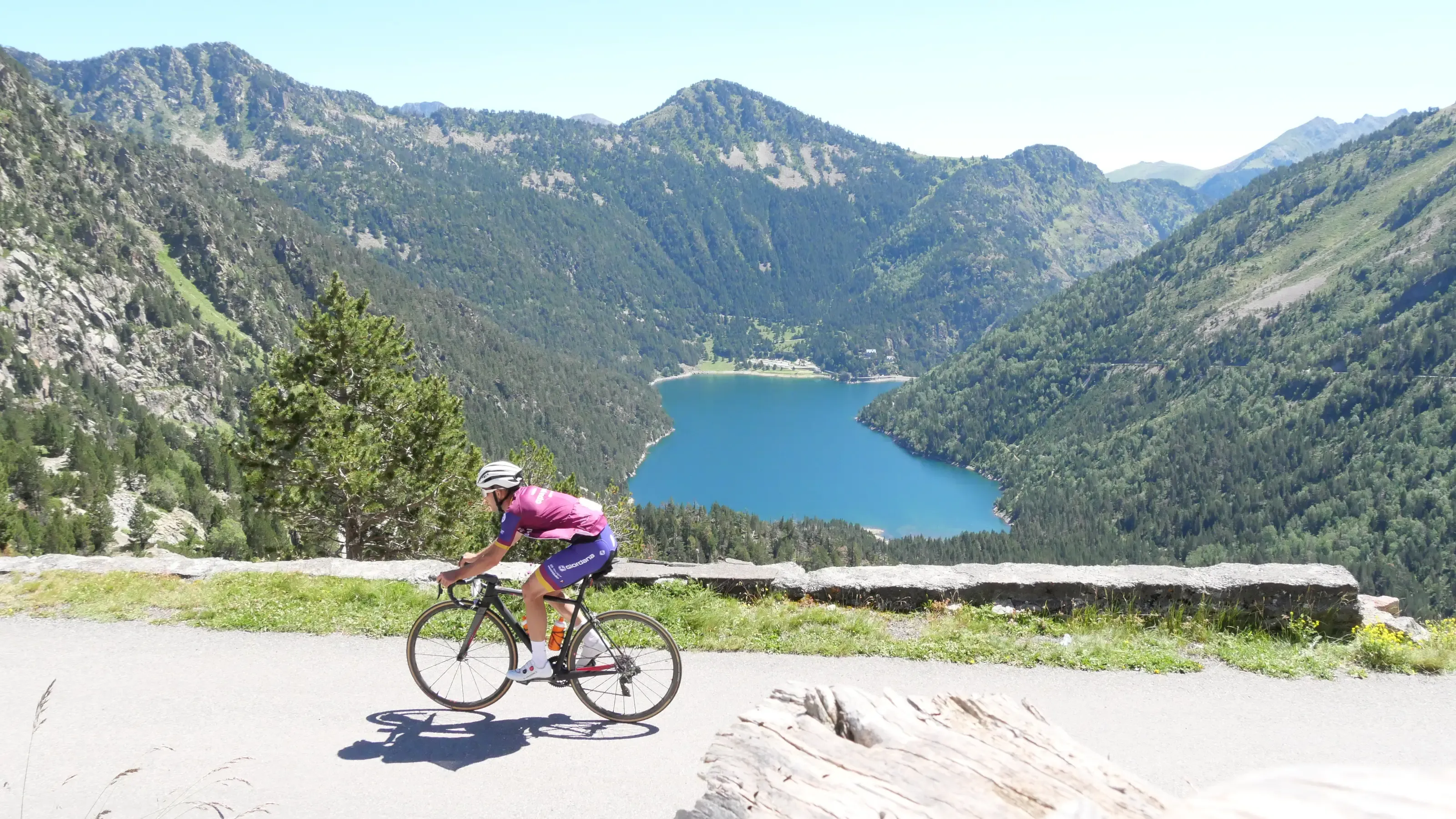 Un cycliste roule sur la route du col de Piau Engaly ensoleillée offrant une vue plongeante sur un lac turquoise au milieu de sommets boisés.